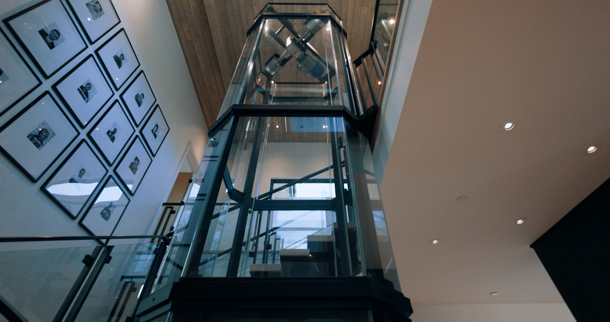 Promontory Model Home Park City Utah — full-height glass residential elevator with black steel frame, gallery wall, and wood-plank ceiling detail