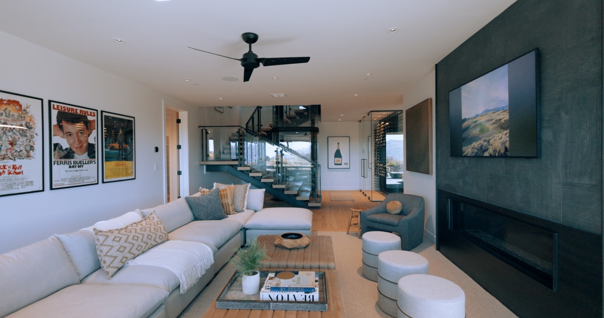 Promontory Model Home Park City Utah — lower-level lounge with L-shaped sectional, dark fireplace wall, glass elevator, and vintage poster art gallery