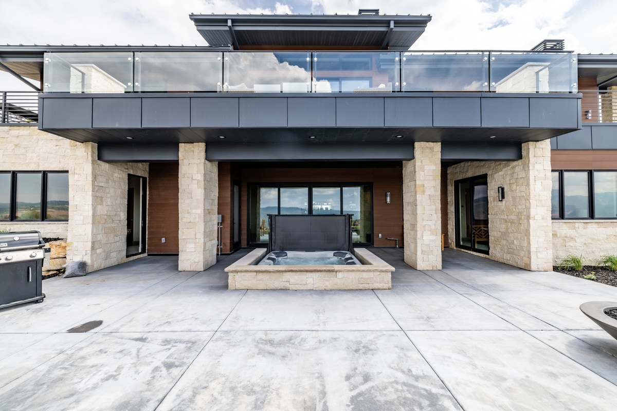 Summit Estates Park City Utah — covered lower patio with stone columns, recessed hot tub, and glass-panel upper deck railing